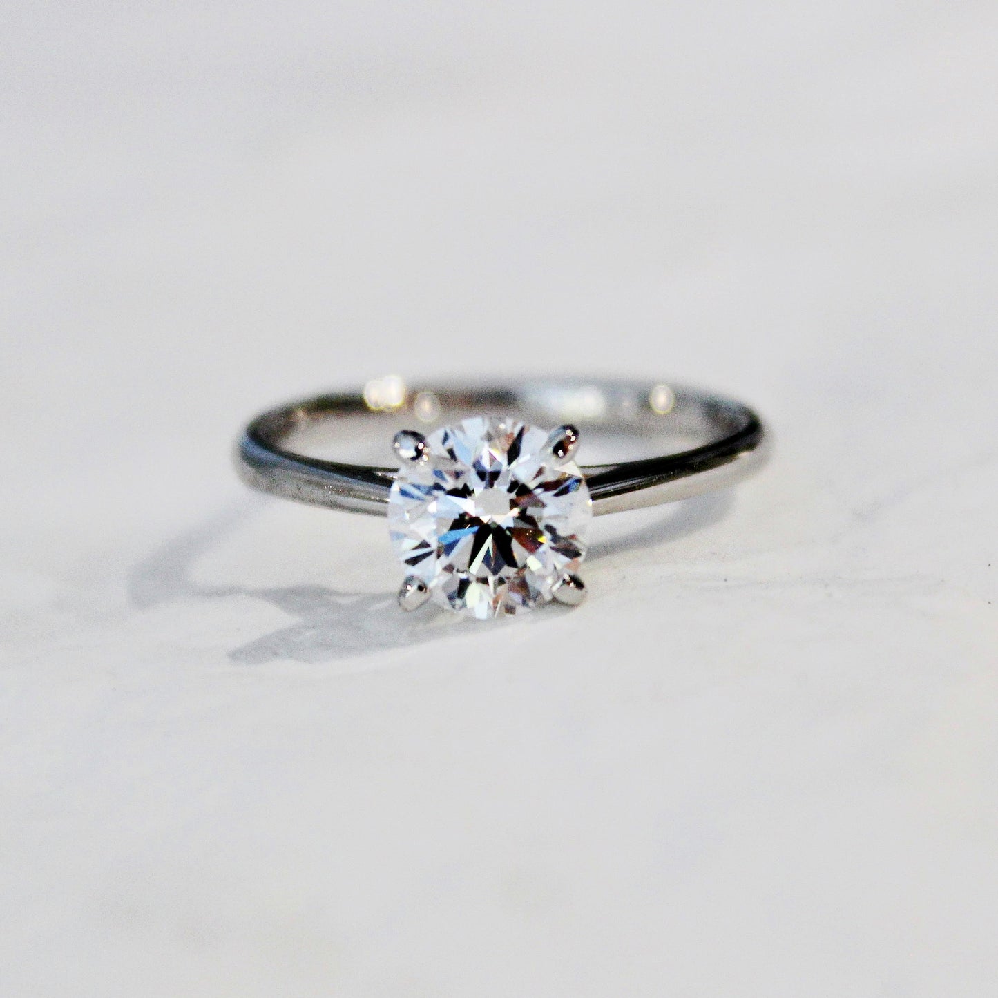 A close-up of a four-prong solitaire engagement ring with a sparkling lab-grown diamond, set in a sleek 18K white gold band, displayed on a white surface.