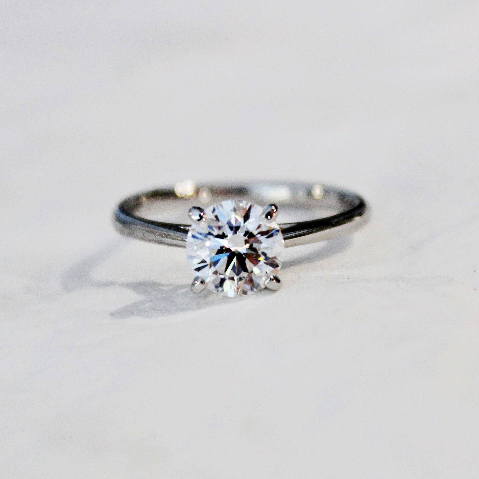A close-up of a four-prong solitaire engagement ring with a sparkling lab-grown diamond, set in a sleek 18K white gold band, displayed on a white surface.