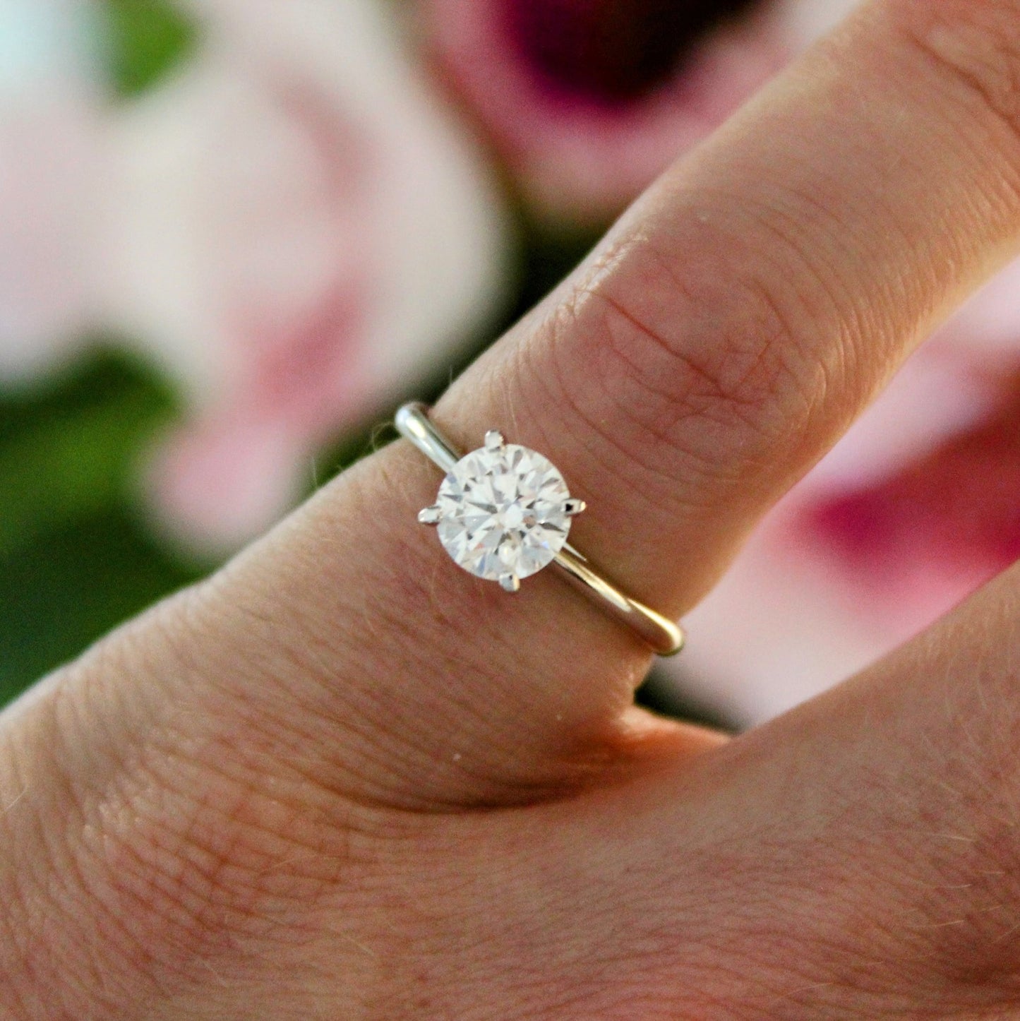 A close-up view of a four-prong solitaire engagement ring on a hand, featuring a sparkling lab-grown diamond set in 18K white gold against a blurred floral background.