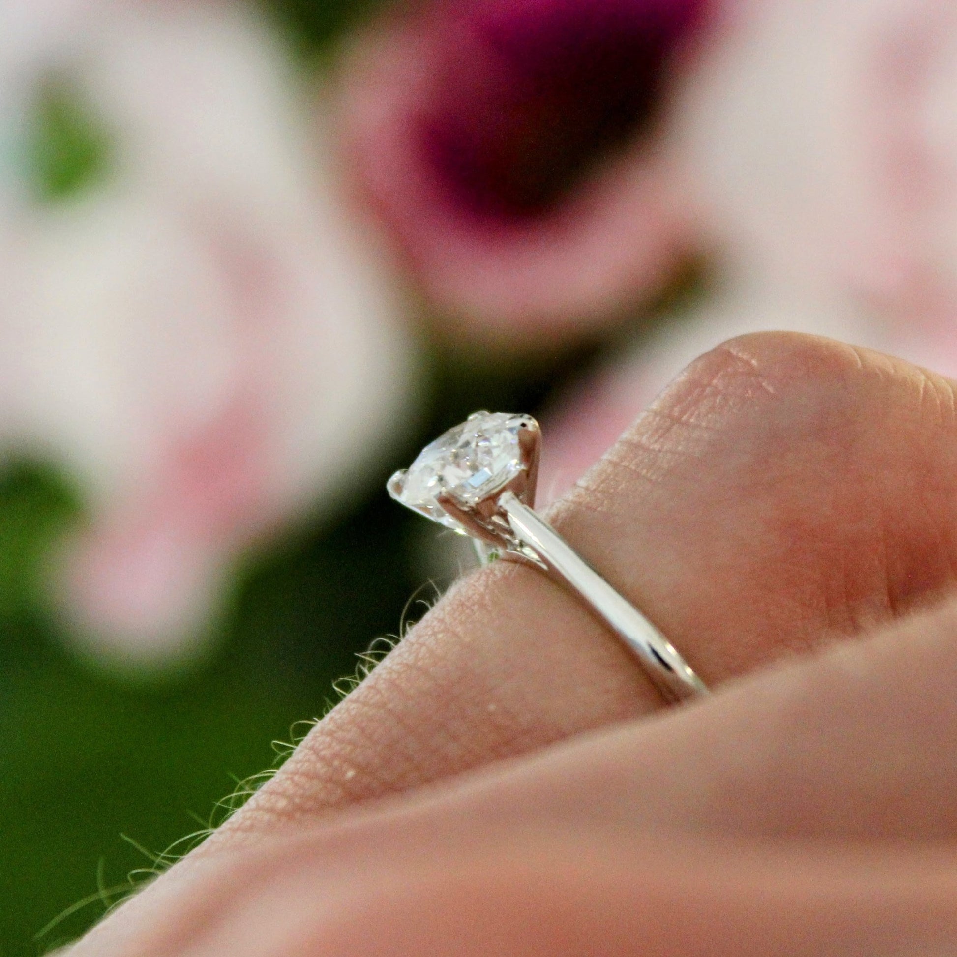 A close-up side view of a four-prong solitaire engagement ring, highlighting the sparkling lab-grown diamond set in a sleek 18K white gold band, with a soft floral background.