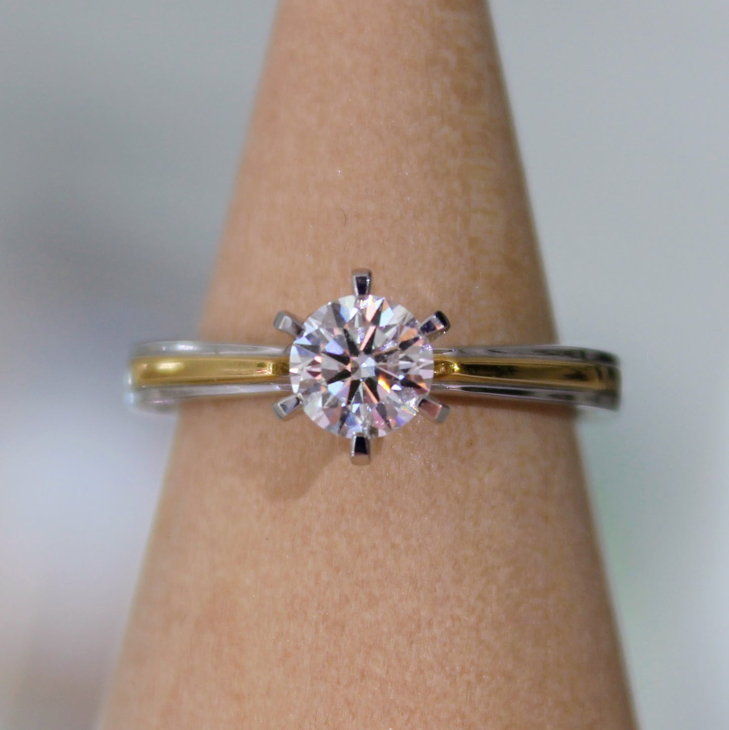A close-up of a two-tone diamond engagement ring displayed on a wooden ring holder, featuring a brilliant solitaire diamond set in 18K solid gold. The perfect blend of luxury and modern design.