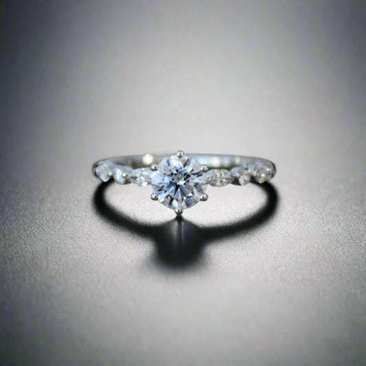 A close-up of a vine-inspired engagement ring in 18K white gold, featuring a round center diamond and marquise-shaped side diamonds, displayed on a stone surface with a soft floral background.